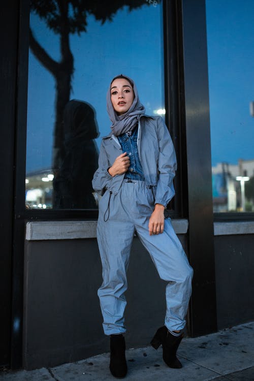 蓝色牛仔夹克和灰色裤子站在玻璃窗口旁边的女人 · 免费素材图片