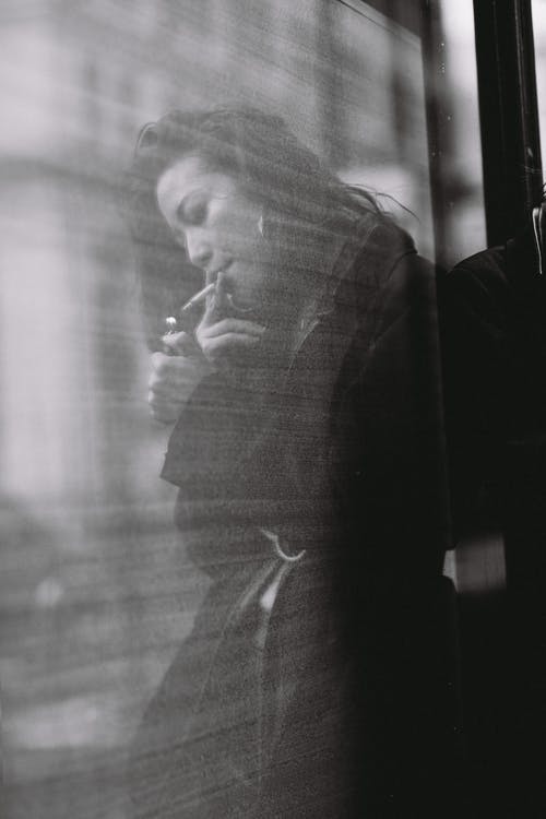 沉思的年轻民族女士吸烟在大街上的玻璃墙附近 · 免费素材图片