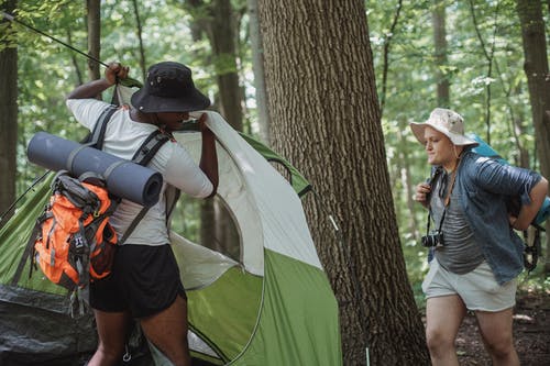 穿短裤和帽子的旅行男人在树林里搭野营帐篷 · 免费素材图片