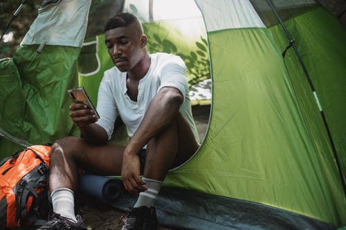 不开心的黑人男子，在帐篷里冲浪智能手机 · 免费素材图片