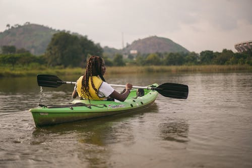 黄色和黑色救生衣的女人在河上骑绿色皮划艇 · 免费素材图片