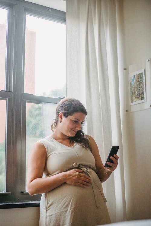 怀孕的女士站在房间里时使用电话 · 免费素材图片