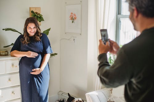 裁剪人怀孕的妻子在室内的智能手机上拍照的人 · 免费素材图片