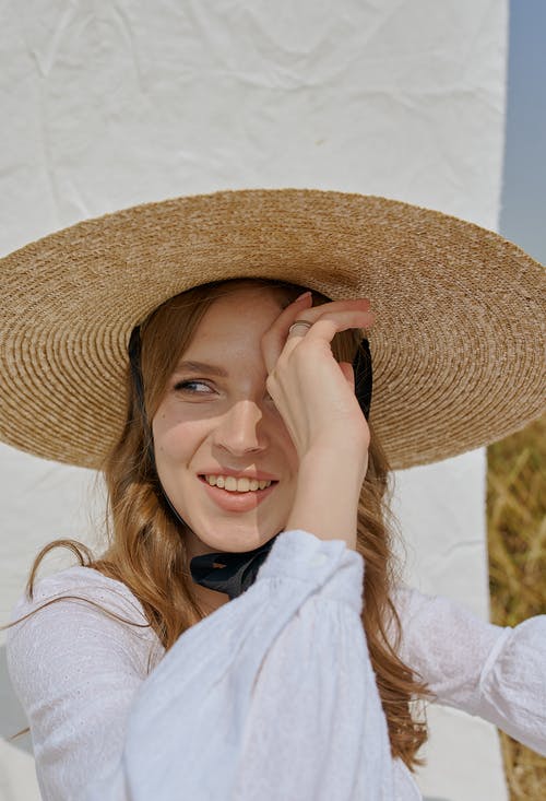 帽子站立在白色板料附近的快乐的妇女在乡下 · 免费素材图片