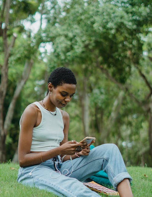 微笑的非洲裔美国年轻女子坐在草地上，使用智能手机 · 免费素材图片