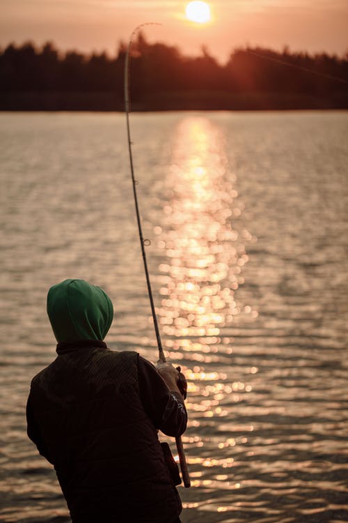 日落期间在海上钓鱼的黑色外套和绿色针织帽的人 · 免费素材图片