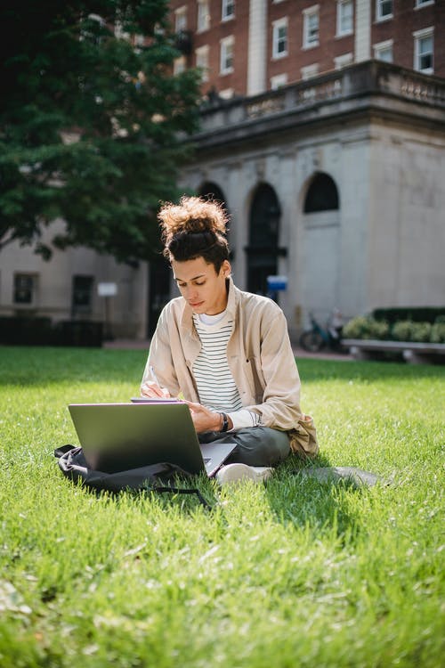 米色的长袖衬衫，坐在绿色的草地上，在使用黑色便携式计算机期间的人 · 免费素材图片