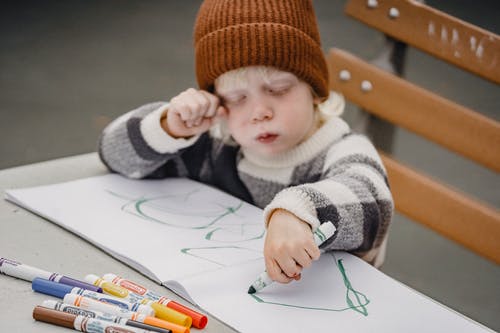 白色和棕色毛衣和橙色编织帽手握一支笔的孩子 · 免费素材图片