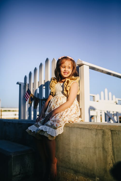坐在混凝土长凳上的白色和棕色花裙子的女孩 · 免费素材图片