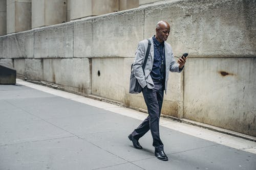 积极时尚的黑人男子，使用智能手机和在人行道上行走 · 免费素材图片