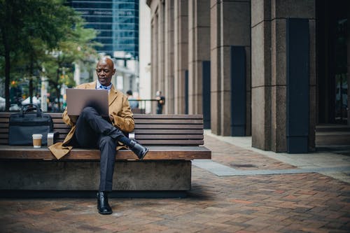 严重的黑人商人，在长凳上的城市街道上使用笔记本电脑 · 免费素材图片