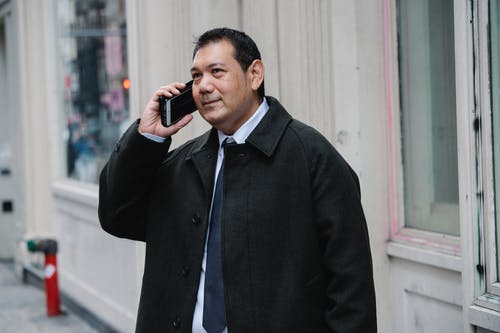 穿着讲究的族裔商人谈老年建筑附近的智能手机 · 免费素材图片