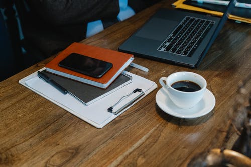 智能手机放在笔记本电脑和工作桌上的咖啡杯附近的记事本上 · 免费素材图片