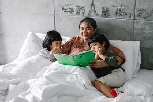 可爱的小亚洲兄弟姐妹躺在床上与微笑的祖母和看书 · 免费素材图片