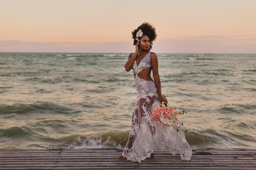 时尚的黑人新娘与鲜花花束走在海洋附近 · 免费素材图片