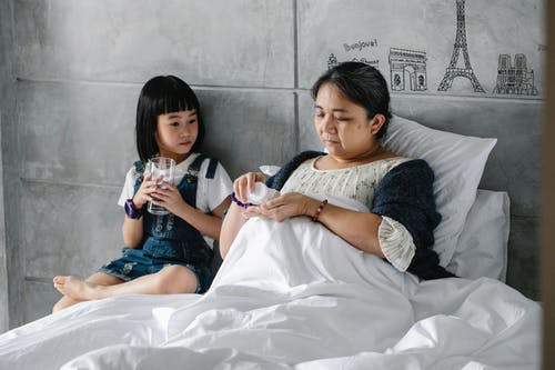 被集中的小亚裔女孩坐在病的祖母饮用的药片附近的床上 · 免费素材图片
