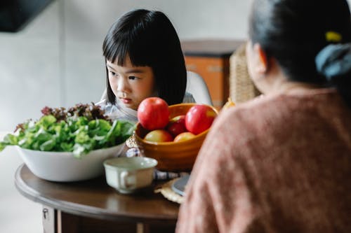 严重的小族裔女孩坐在餐桌旁与母亲在午餐期间和视线 · 免费素材图片