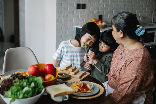 积极的小族裔男孩喂养妹妹在和祖母一起吃午饭的时候 · 免费素材图片