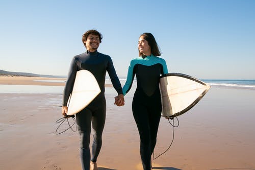 2名穿着蓝色和黑色潜水衣的妇女在海滩上举行白色冲浪板 · 免费素材图片