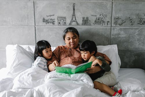 可爱的小民族兄弟姐妹躺在床上和祖母一起读书 · 免费素材图片
