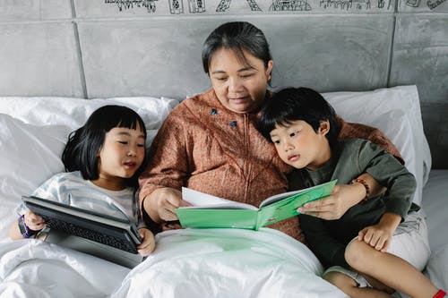 族裔祖母与好奇的孩子躺在床上，有趣的书和平板电脑 · 免费素材图片