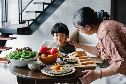 可爱的亚洲男孩在厨房里细心的奶奶附近吃面食 · 免费素材图片