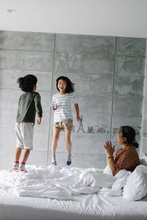 兴奋的族裔小孩子在拍手祖母附近的床上跳 · 免费素材图片