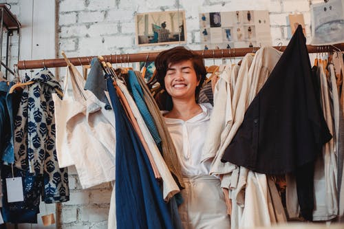 站立在垂悬的衣裳中的快乐的亚裔女性顾客在商店和微笑中 · 免费素材图片