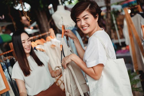 站立与在挂衣架的礼服的快乐的亚裔妇女在市场上 · 免费素材图片