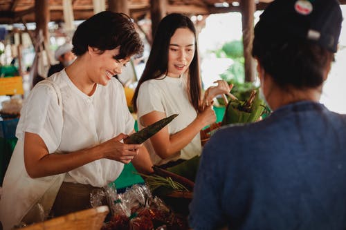 微笑族裔购物者在市场中无法识别的卖方附近选择蔬菜 · 免费素材图片