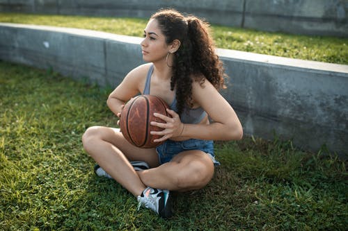 蓝色牛仔短裤的女人坐在绿草地上举行篮球 · 免费素材图片