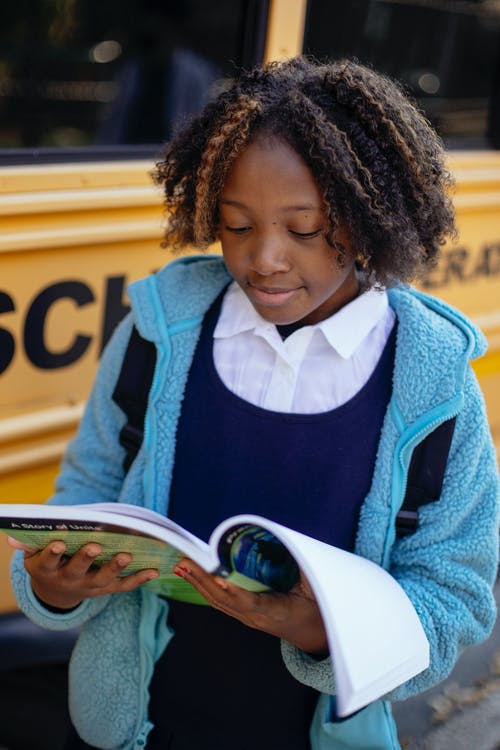 正面黑人女孩读书课本在校车附近 · 免费素材图片