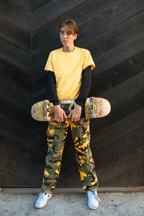 黄色v领毛衣和迷彩裤，拿着棕色和黑色滑板的人 · 免费素材图片