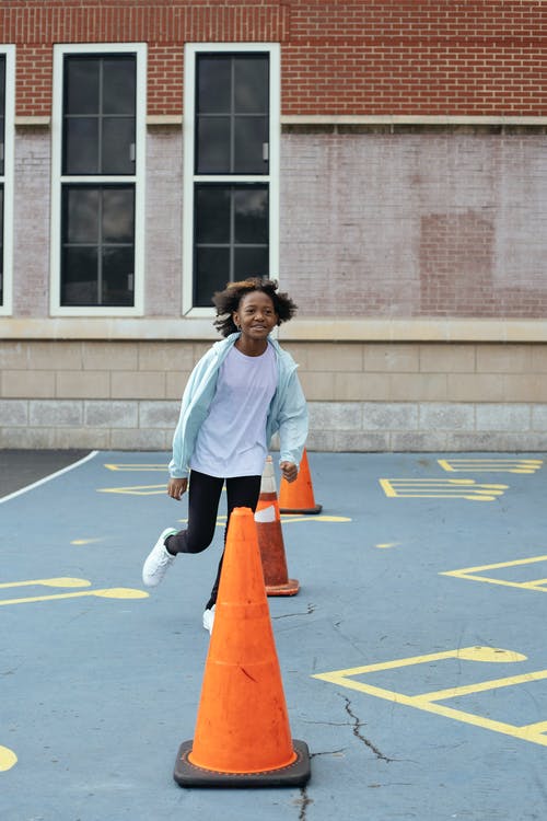 快乐的黑人女孩，在学校附近的操场上的红色锥体周围跑 · 免费素材图片