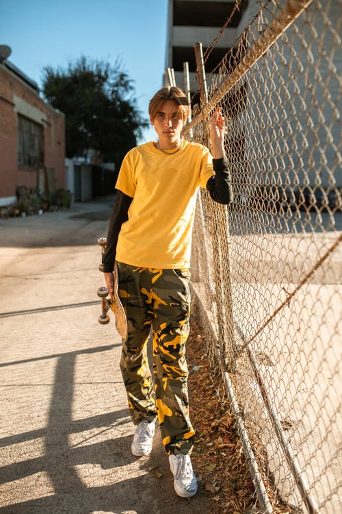 穿着黄色圆领t恤和棕色迷彩裤的人倚在灰色金属栅栏上 · 免费素材图片