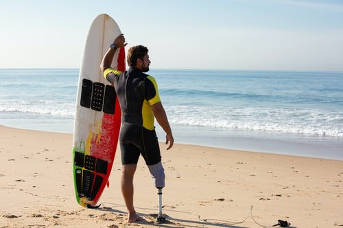 男性的截肢者冲浪者站在海滩上的冲浪板 · 免费素材图片