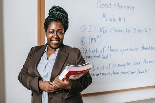 性格开朗的黑人女老师与作业簿站在白板附近 · 免费素材图片