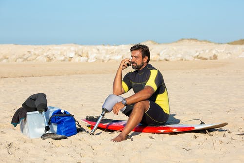 被截肢者男性冲浪者，坐在冲浪板上，和谈智能手机 · 免费素材图片