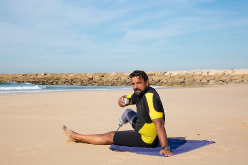 男性截肢者冲浪者在沙滩上休息，喝些热水瓶 · 免费素材图片