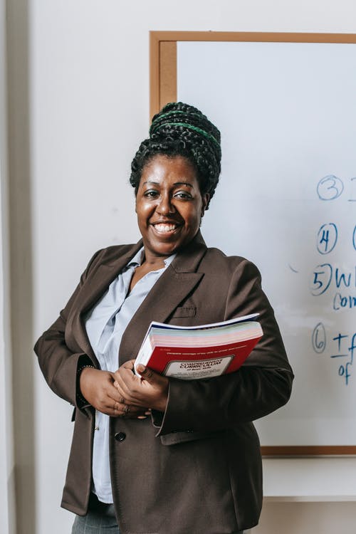 站立与作业簿的愉快的黑人女老师在白板附近 · 免费素材图片