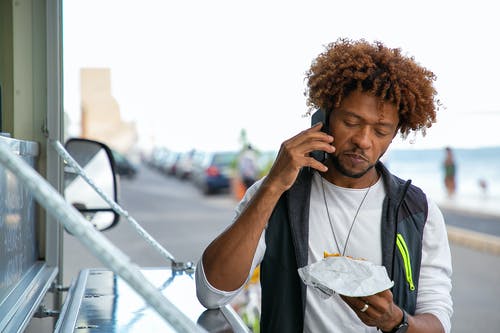 黑人在街上吃汉堡时谈智能手机 · 免费素材图片