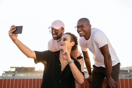 快乐年轻的多种族男性朋友显示胜利手势和采取自拍照在智能手机上 · 免费素材图片