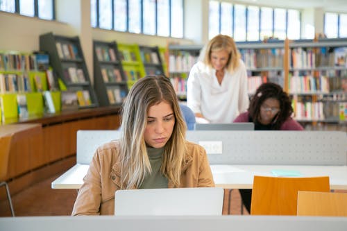 严重的年轻女子，与不同的组员在图书馆的笔记本电脑上工作 · 免费素材图片