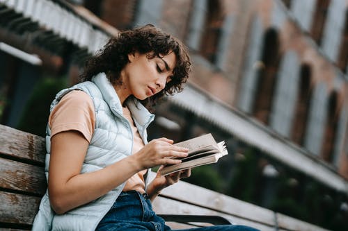 白衬衫和蓝色牛仔牛仔裤，坐在黑色的金属长椅上看书的女人 · 免费素材图片