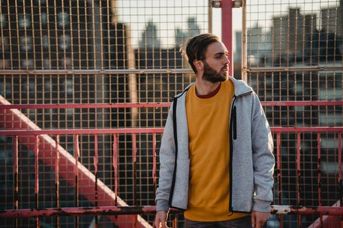 身穿橙色圆领衬衫和灰色夹克站在红色金属栅栏旁边的人 · 免费素材图片