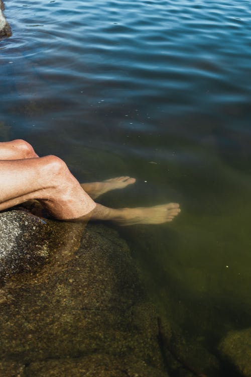 无法认出的人在岩石湖岸上休息时浸入水中的脚 · 免费素材图片