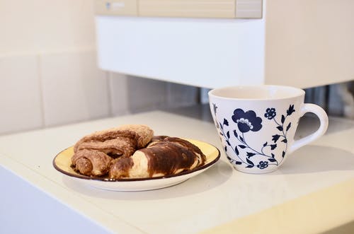 白色陶瓷板上黑面包旁边的白色和蓝色花卉陶瓷杯 · 免费素材图片