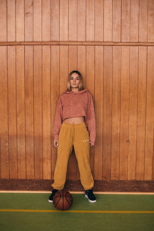 棕色长袖衬衫和黄色的裤子，站在棕色木墙旁边的女人 · 免费素材图片