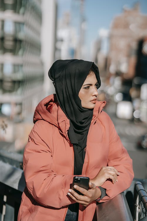 黑色的头巾和橙色外套拿着智能手机的女人 · 免费素材图片