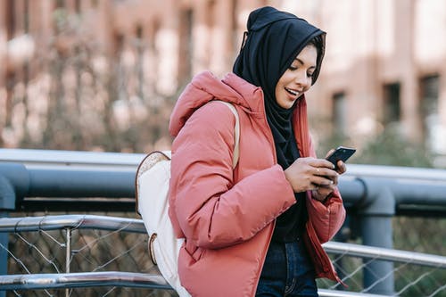 黑色的头巾和持有智能手机的粉红色外套的女人 · 免费素材图片